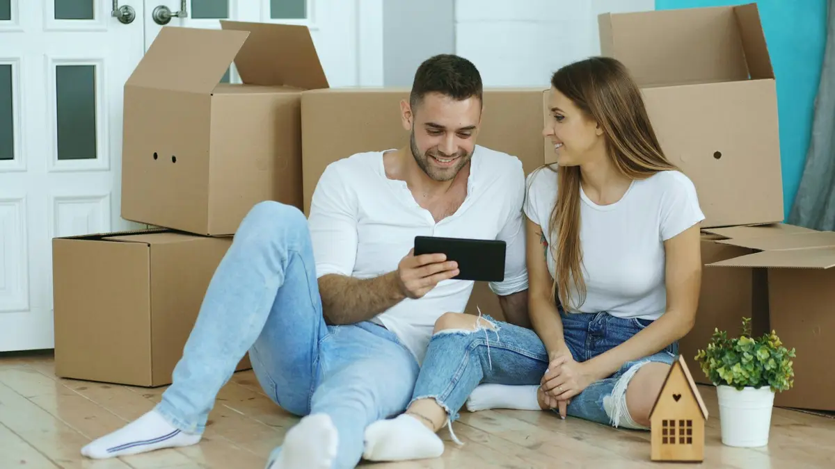 Casal sentado no chão entre caixas de mudança, olhando imóveis em um tablet